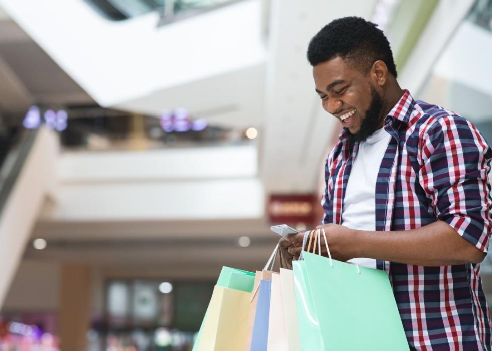 A smiling man in a shopping mall is holding several colorful shopping bags in one hand while looking at his smartphone.