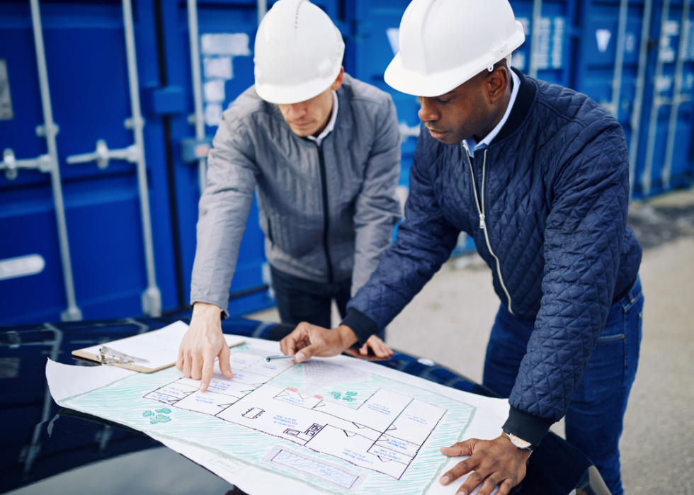 Two people in hard hats standing near shipping containers look at a large blueprint.