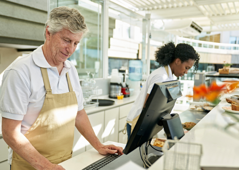 An older man wearing an apron behind a counter in a cafe looking at the computer in front of him. African American woman in the background is leaning towards the counter in front of her. 