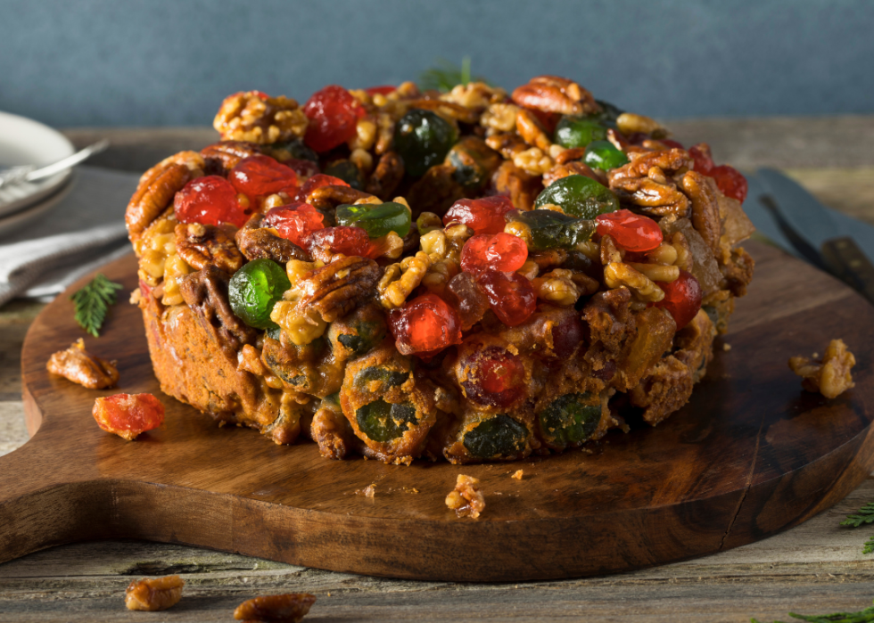 A round brown cake on a wooden cutting board with sprinkled nuts and candied fruit.