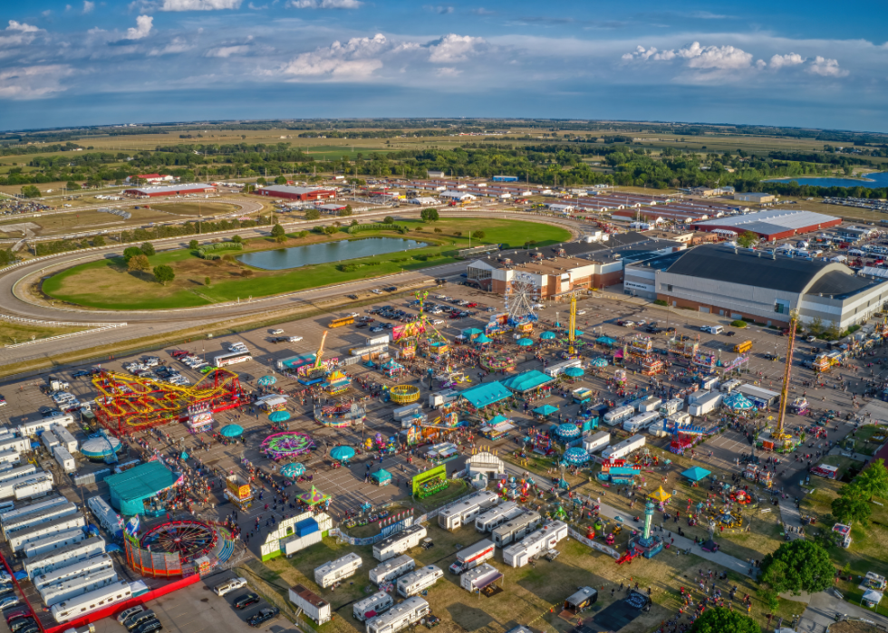 Aerial view of colorful rides with a large ferris wheel, RVs, and crowds of people attending state fair. 