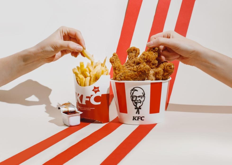 Two hands reaching for fries and chicken out of the KFC bucket filled with fried chicken and french fries. 