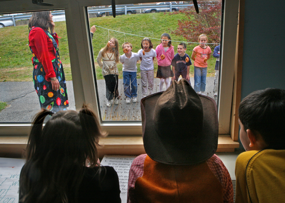 A group of elementary school students watch from a window as other students bob for donuts during recess on Halloween. 