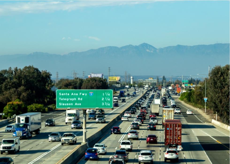 A multi-lane highway with heavy traffic. The road is divided by a concrete barrier, and there are several overpasses visible in the background. The sky is clear blue, and there are mountains in the distance.