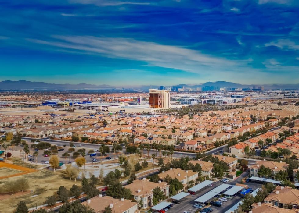  An aerial view of  a sprawling suburban landscape in a sunny climate. The image features a vast expanse of residential homes with a variety of styles, including single-family houses and townhomes. 