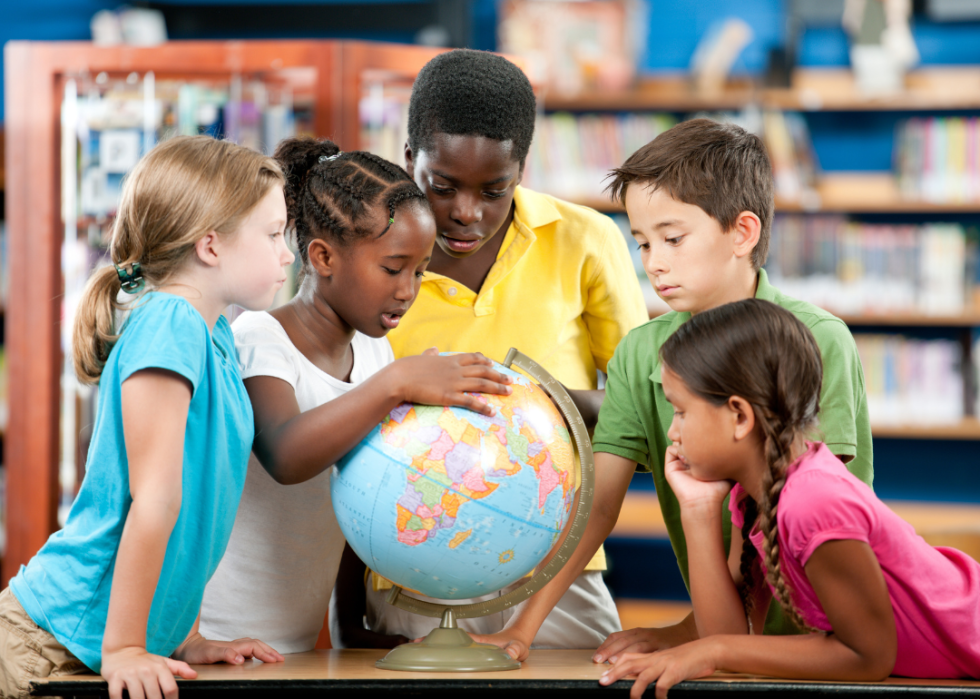 A group of five young children gathered around a globe in a classroom or library setting. The children, who are of different ethnic backgrounds, are all focused on the globe. 