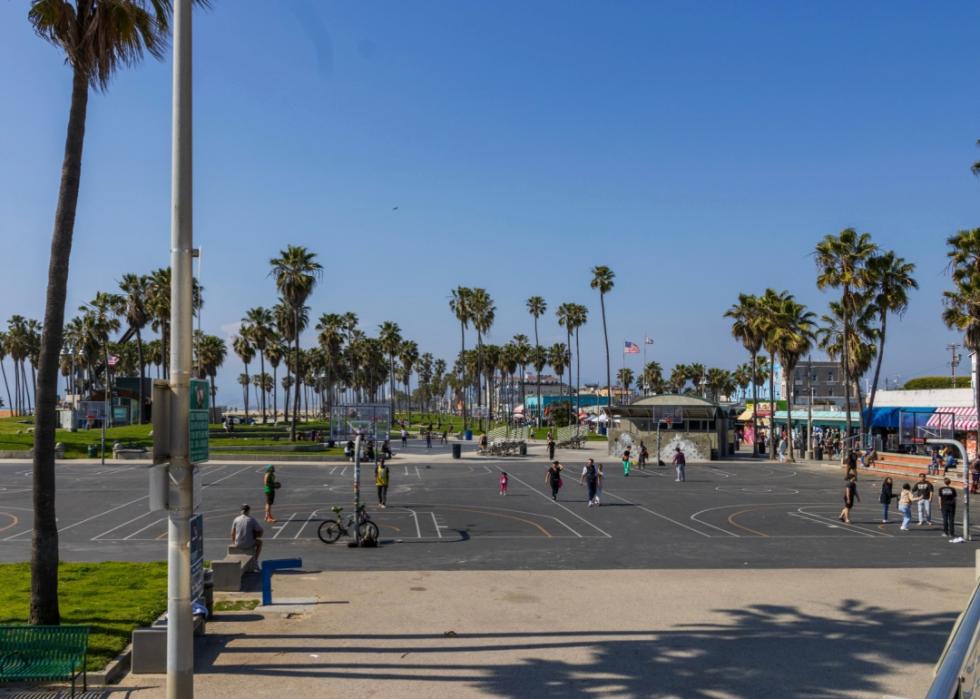 A basketball court with people playing. The court is surrounded by buildings and palm trees. The sky is blue and sunny. There are a few people watching the game.