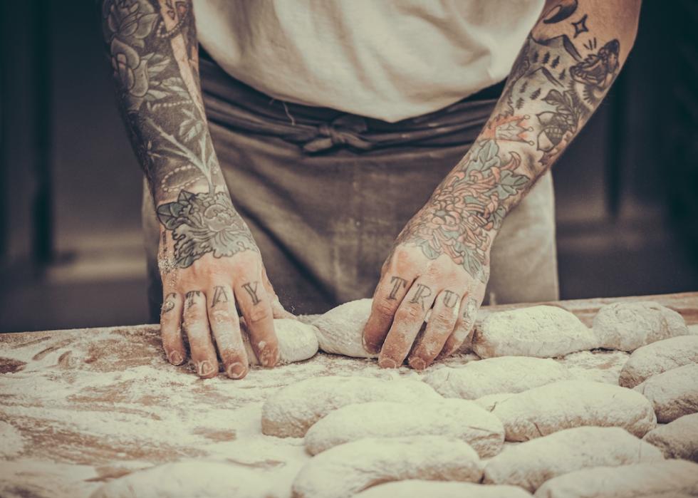 Baker with tattoos puts raw dough buns on the baking tray in the bakery.