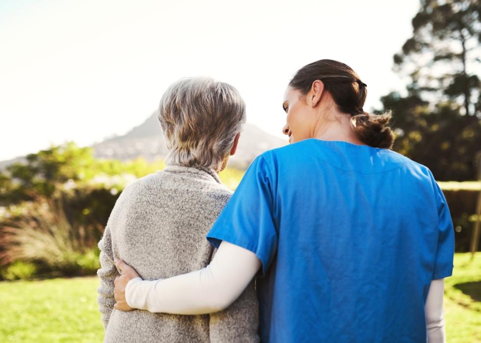 Rear view of a mature patient with a home health aide walking outdoors.