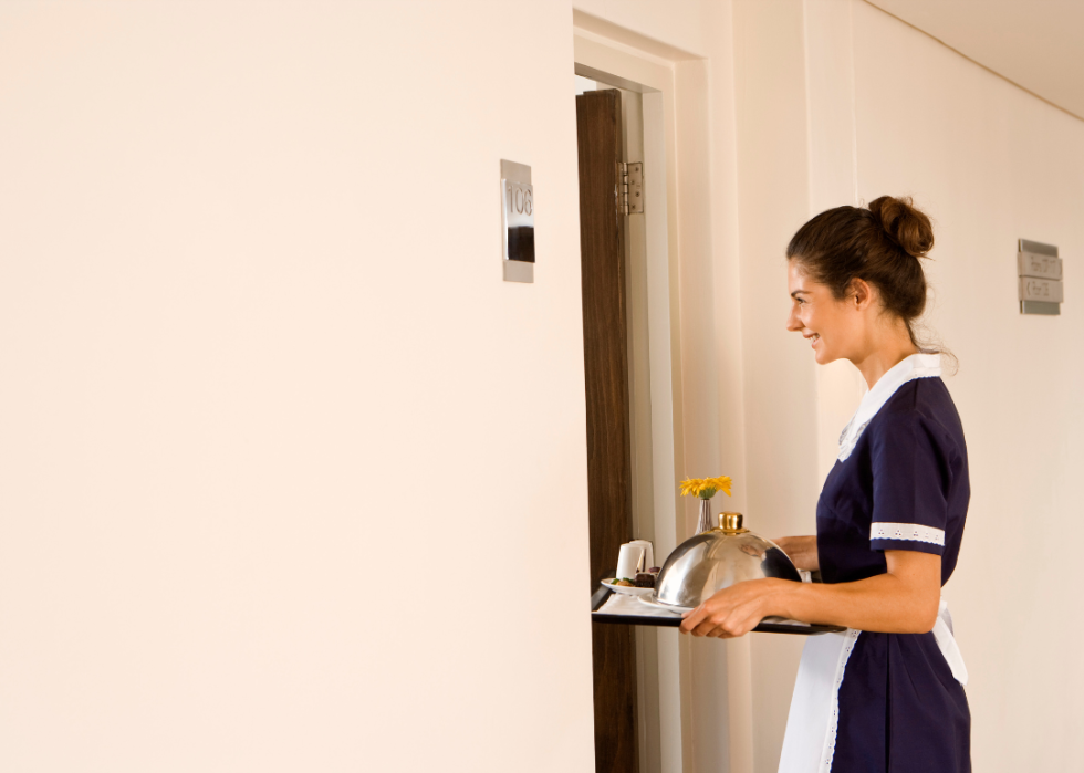 A smiling maid holding a tray standing in front of a room.