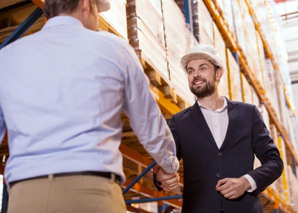 Two men wearing helmets shaking hands while meeting each other in a warehouse.