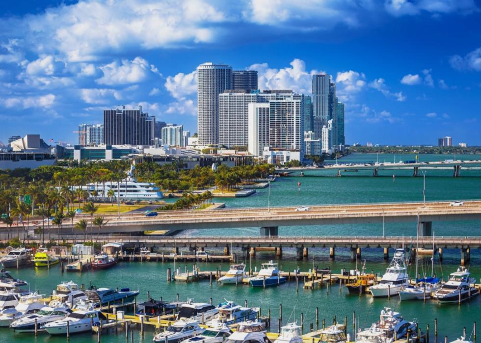 A coastal city with a stunning skyline. The photo features a bustling marina filled with yachts and boats, with a bridge spanning the waterway. In the background, towering skyscrapers 