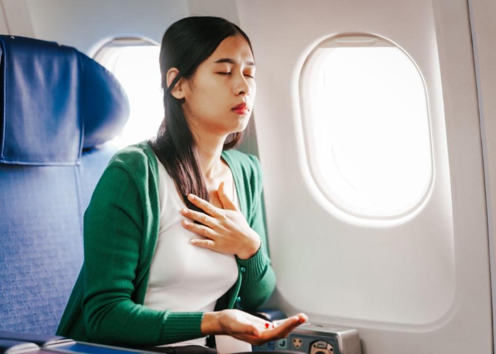 A woman is sitting in her seat as an airplane passenger, closing her eyes with her hand to her chest and other hand holding pills.