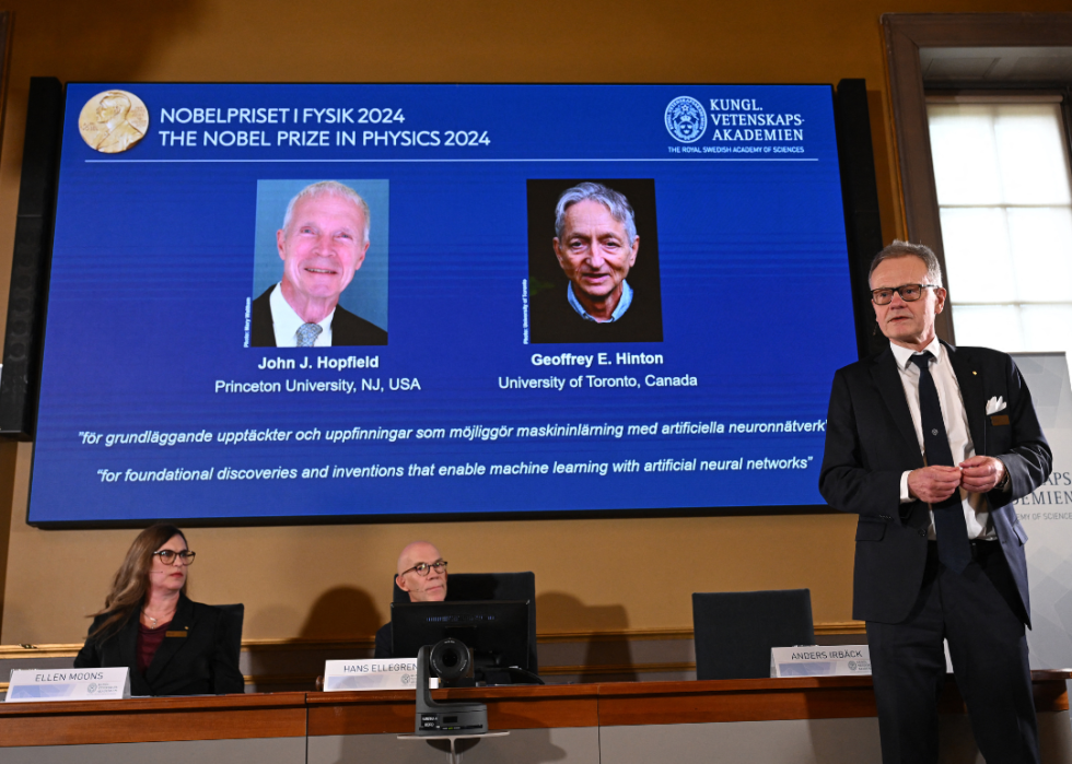 A large screen mounted on a wall behind several committee members shows the laureates of the 2024 Nobel Prize in Physics, John J. Hopfield and Geoffrey E. Hinton.