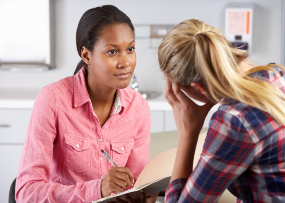 A teenager with her back facing us, resting her head on her hand, and sitting across from a nurse with a clip board in an office.