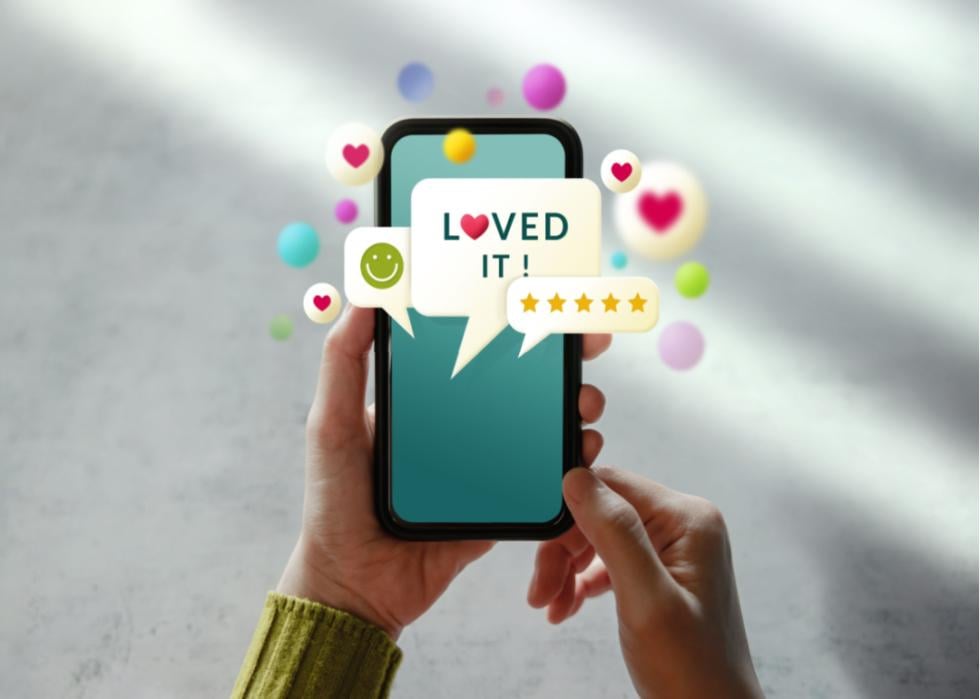 A photo illustration of a close up of hands holding a mobile phone with heart, smiling, and speech bubble emojis floating around it. 