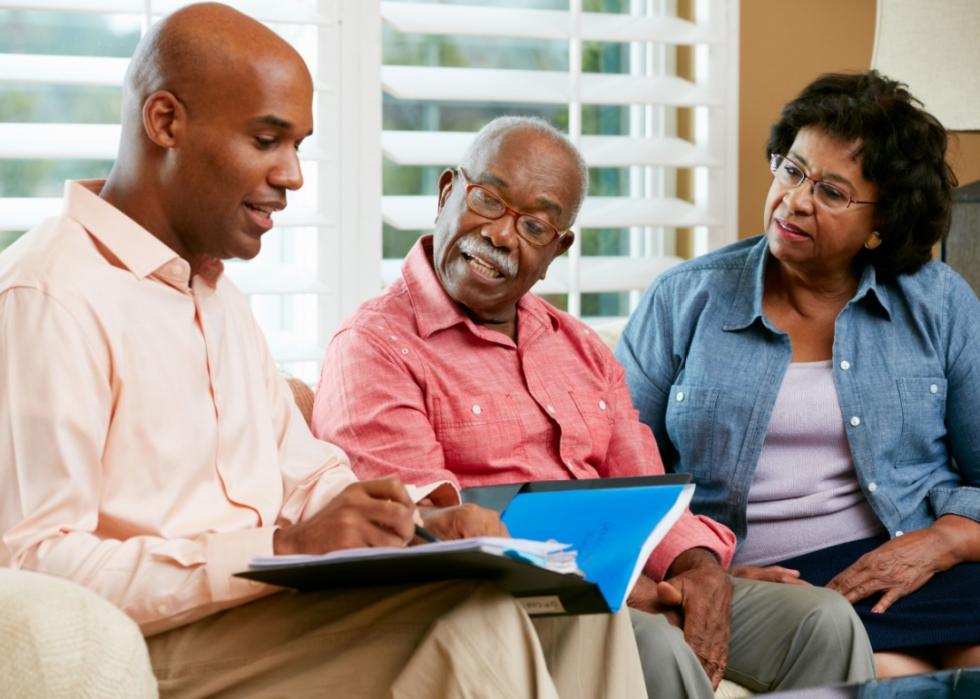 A young Black man sitting on a couch with a folder with papers on his lap next to an older Black couple looking at the folder with him.