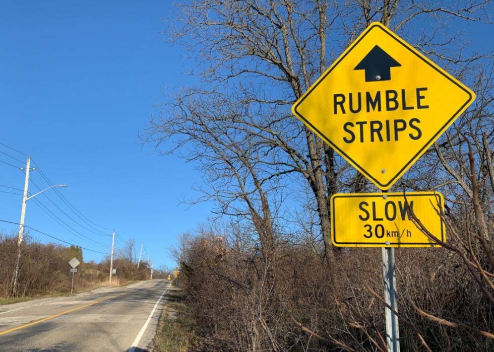 Yellow and black road sign warning of rumble strips ahead on a crumbling, urban road. Series of raised strips seen in the background.