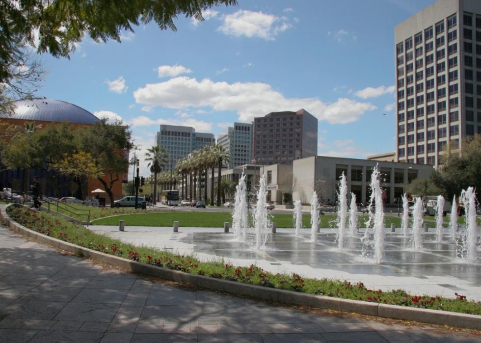 A city park with a fountain. In the foreground, there is a paved area with a few steps leading down to a grassy area. Palm trees and tall building in the background.