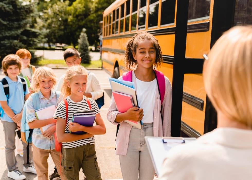 A line of schoolkids with backpacks some holding notebooks standing by schoolbus.