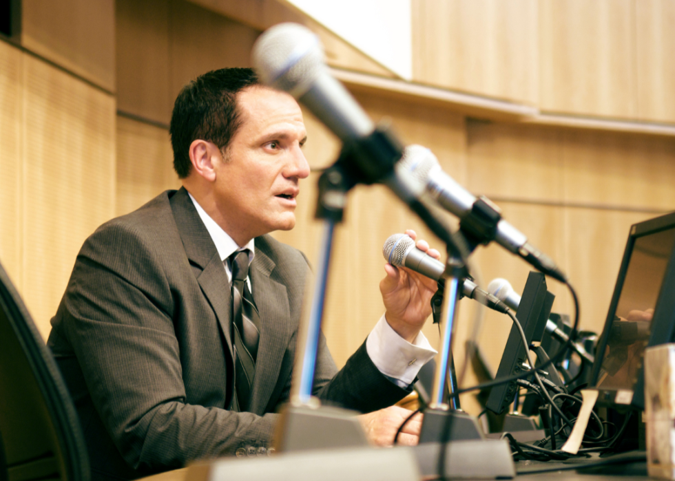 A senator sits at a table in front of microphones. 