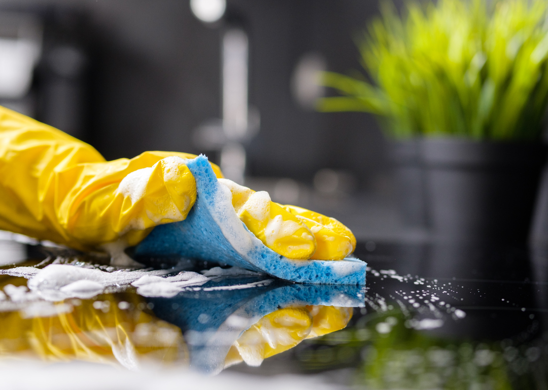 A hand with a yellow rubber glove cleaning a surface with a sponge.