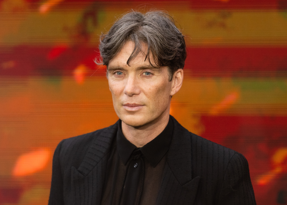 Cillian Murphy in a black suit in front of a red and orange striped background.