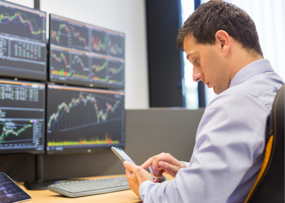 A stockbroker looks at his phone while he sits in front of four computer screens and a tablet. 
