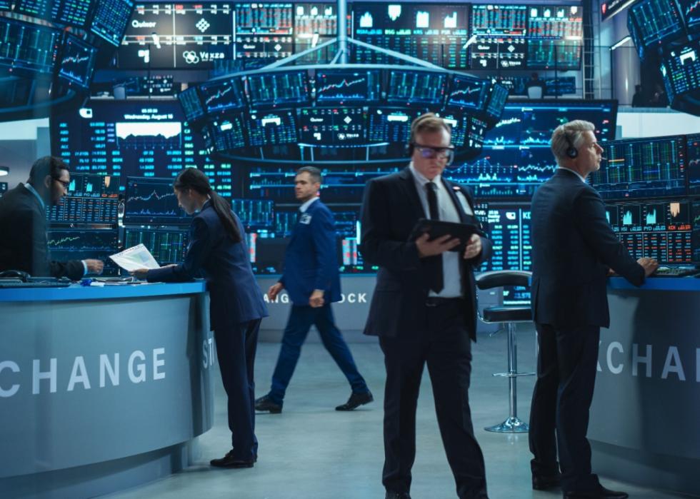 A floor of the stock market with a large wall full of screens showing graphs. Five people are seen in the foreground. A woman showing a piece of paper to a man at a desk as man in a suit walks by. A man with a tablet and headpiece stands in the center while the man on the right is focusing on the screen in front of him.