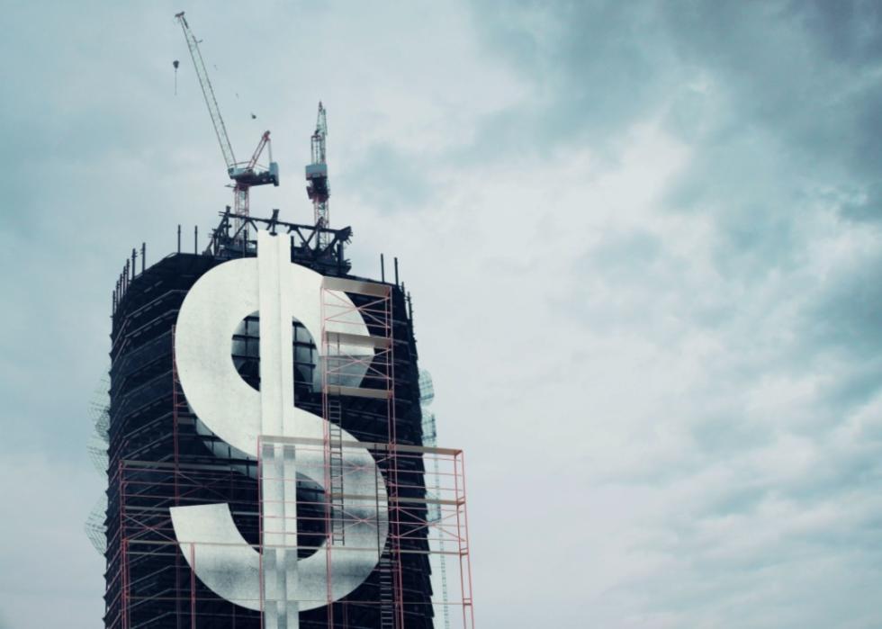 A large buillding under construction, with a prominent dollar sign ($) symbol at the forefront. The building is surrounded by scaffolding, and several cranes are positioned at the top. 