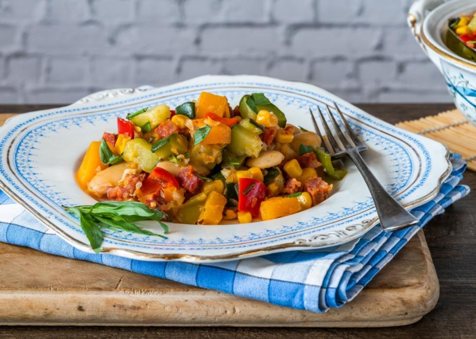 A plate of vegetables, butter beans, butternut squash, sweetcorn and pieces of sausage. The plate sits on top of blue kitchen towel and cutting wooden board. 