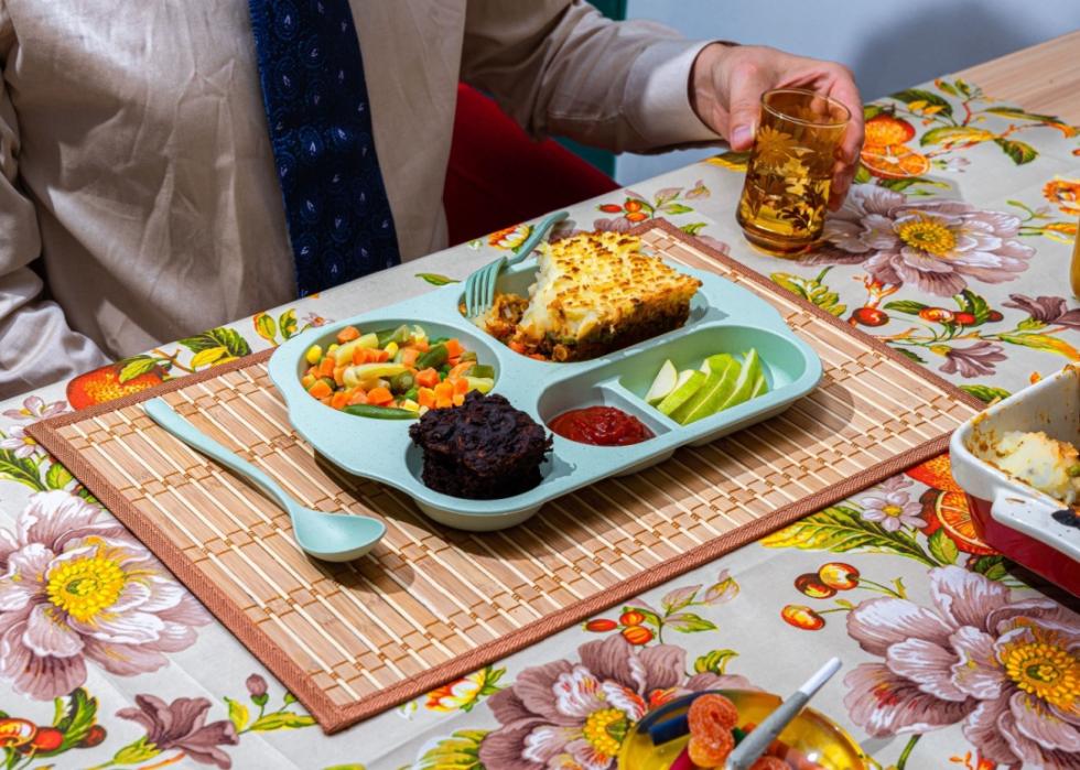 A man sitting in front of the plastic blue tray with a meal compartmentalized into sections with mashed potatoes and ground meat, sliced green apple, mixed veggies and a muffin. 