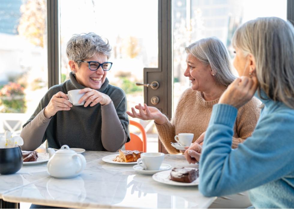 Three older smiling women sitting around a table with white cups, saucers, and a teapot in a bright, casual setting, likely a café. 