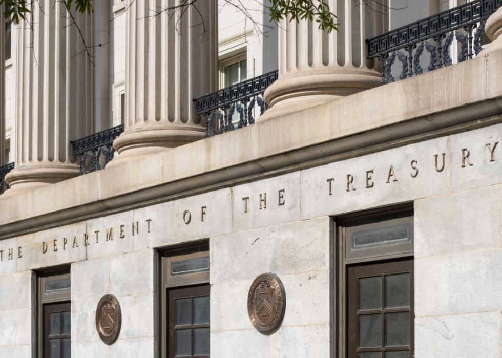 A portion of the facade of a building. The building is made of stone and features columns with ornate capitals. The words “The Department of Treasury” are inscribed in large letters above the entrance.