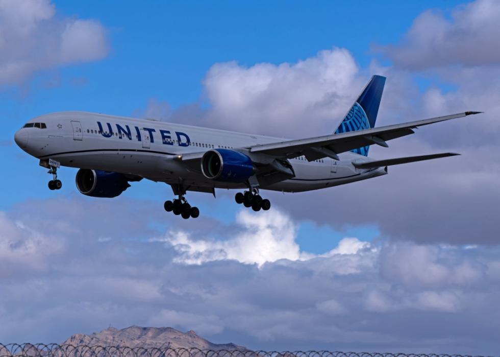 United Airlines airplane in flight. The United Airlines logo and branding are clearly visible on the aircraft's body and tail. The background shows a partly cloudy sky with mountains visible in the distance.