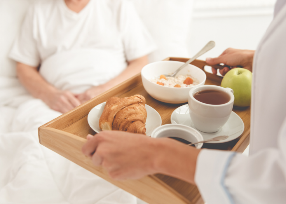 A person serving someone sitting in a bed a tray of food, including an apple, a croissant, a bowl of porridge, and a cup of tea or coffee.