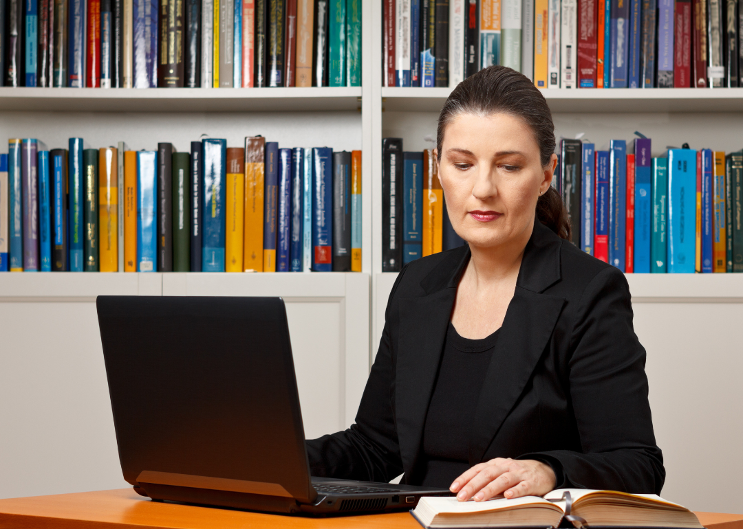A woman with a laptop sits in front of a bookcase.