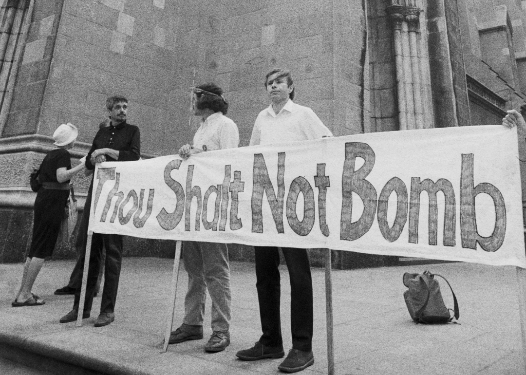 Reverend Daniel Berrigan (wearing black) and others standing behind a "Thou Shalt Not Bomb" sign at St. Patrick's Cathedral in NYC in protest of US bombings in Cambodia.