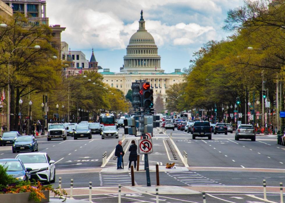 A large building with the white dome in center.  The street in the foreground is busy with traffic, featuring multiple lanes filled with cars, and traffic lights are visible in the middle of the road. On either side, trees line the street. 