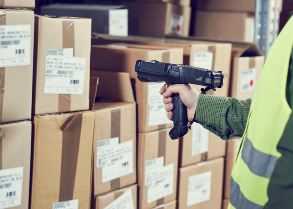 A close-up of a hand holding a barcode scanner pointing to the pile of boxes.