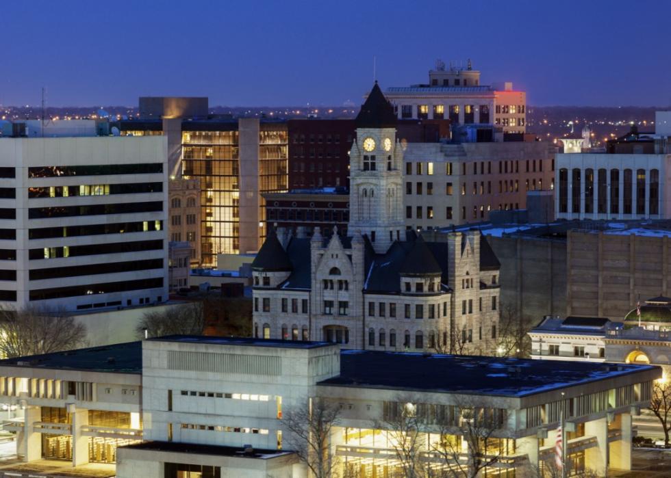 A cityscape at night, centered on a grand, ornate building with a prominent clock tower. The surrounding buildings are a mix of modern and older architectural styles.