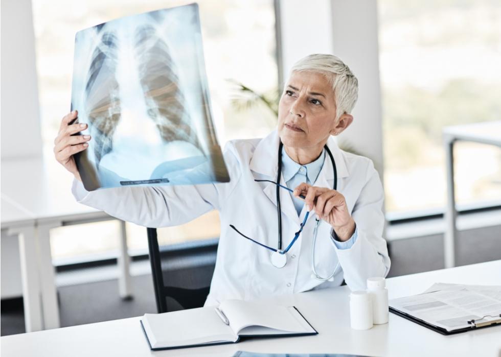 An older female doctor is looking focused at lungs x-rays she is holding up.