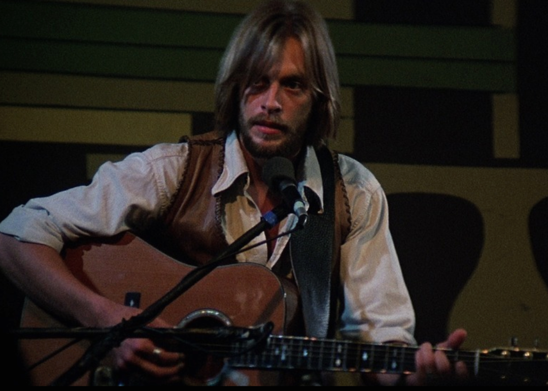A man with long hair plays the guitar in a long sleeved shirt and leather vest.