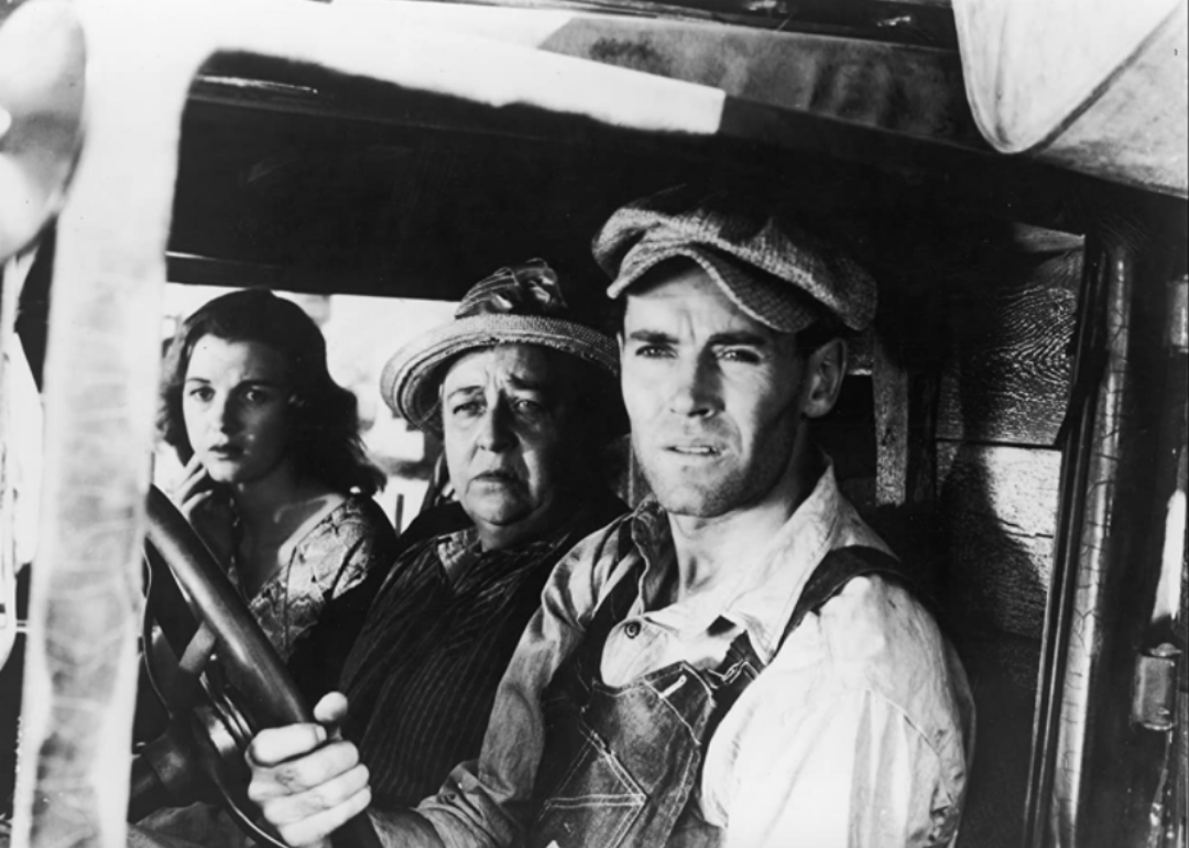 A man drives an old car with an older woman and a girl in the passenger seat, all looking very serious.