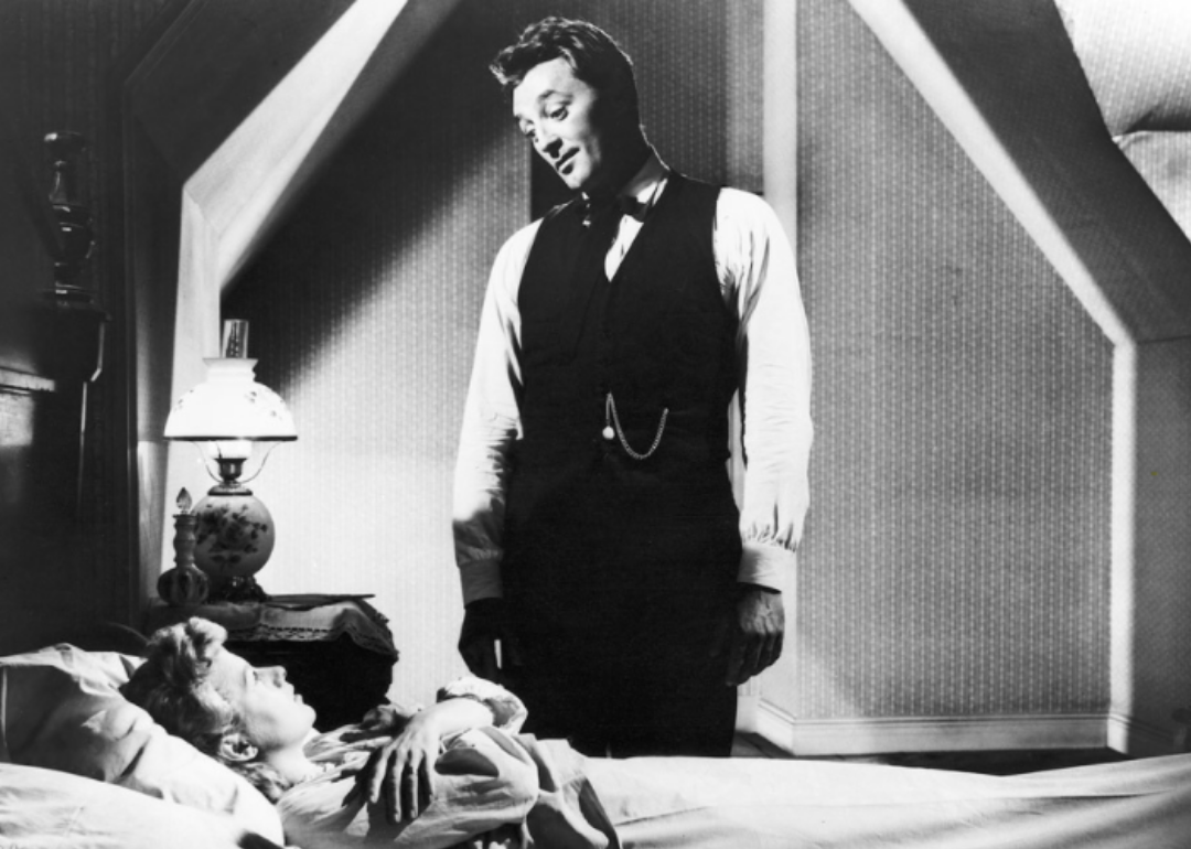 A man stands over a woman in bed looking at him with her arms crossed.