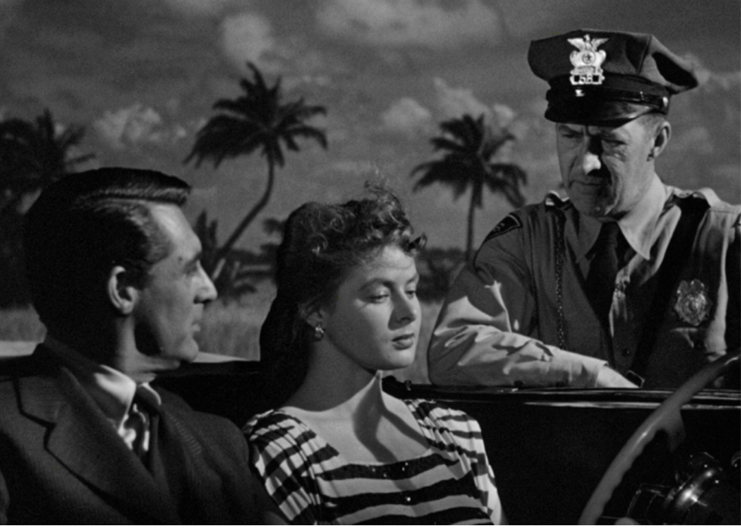 Ingrid Bergman and Cary Grant pulled over by a police officer in their car with palm trees in the background.