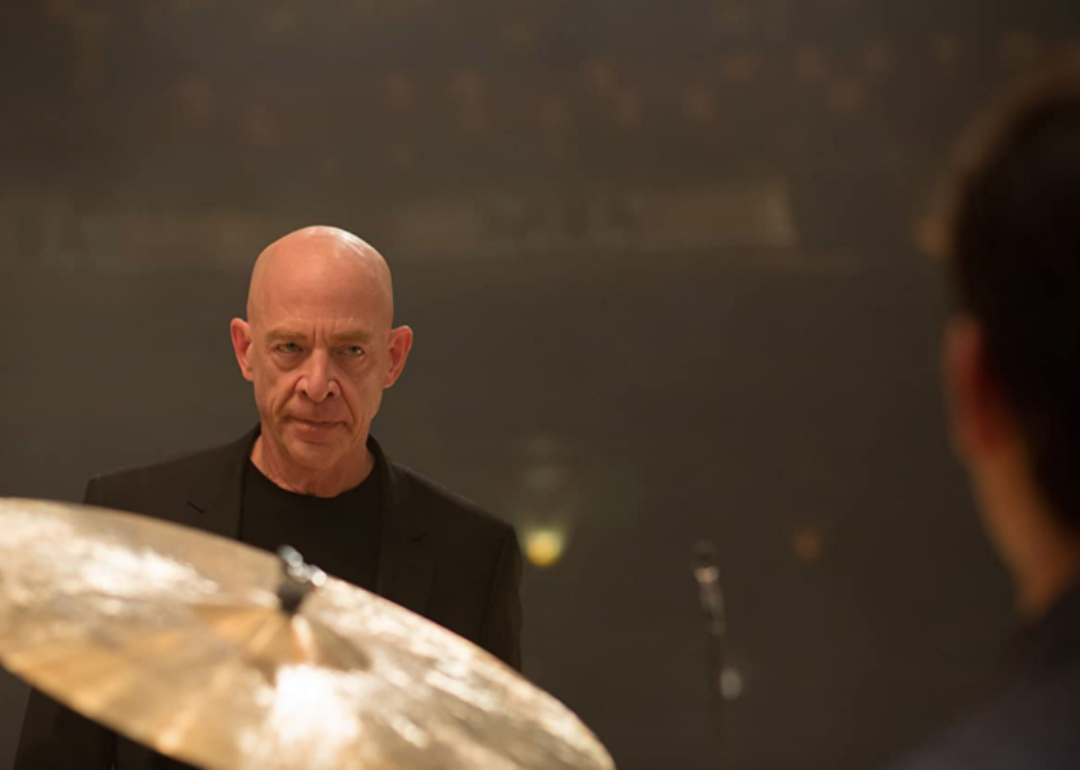 J.K. Simmons with a serious look on his face looking at someone drumming.