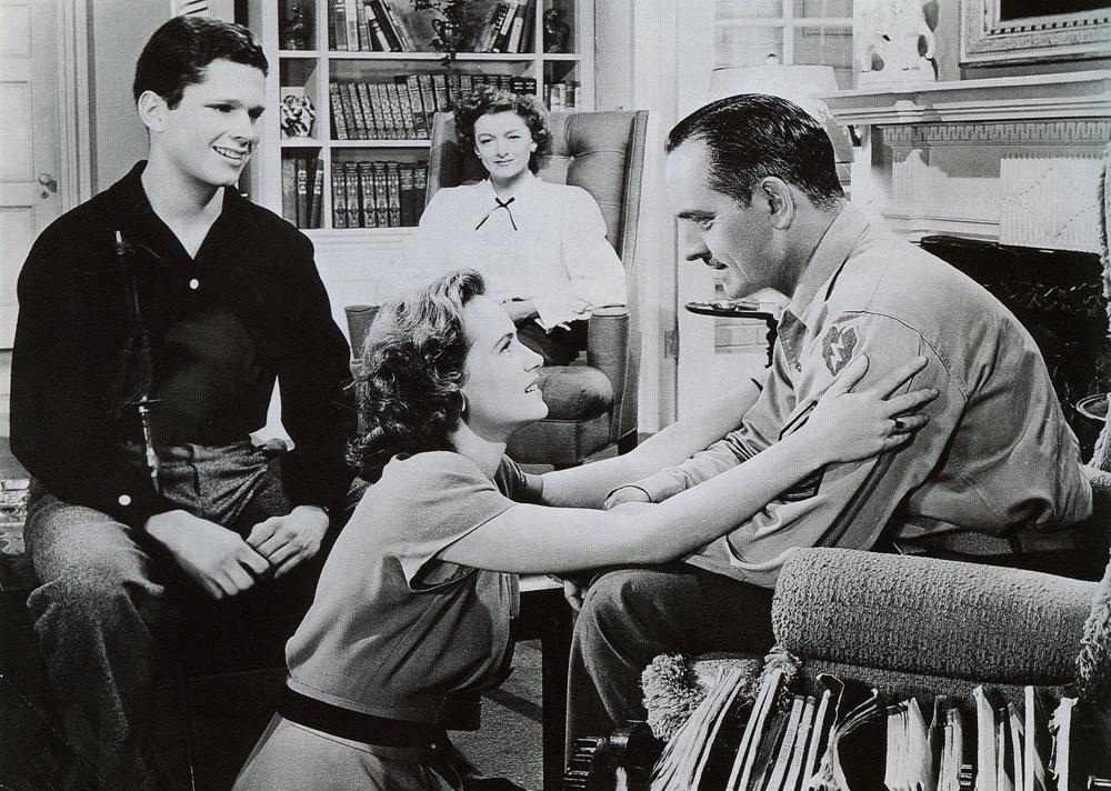 A family sitting in a living room with a woman kneeling and smiling up at a man sitting in a chair.