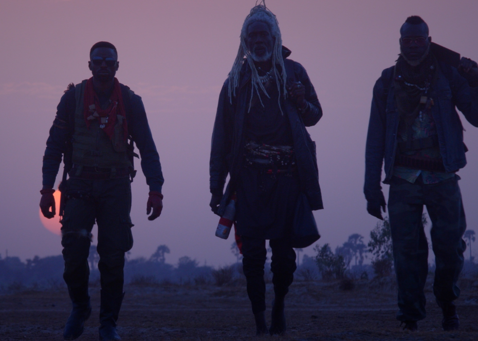 Three men dressed like warriors walk with a sunset and pink sky in the background.