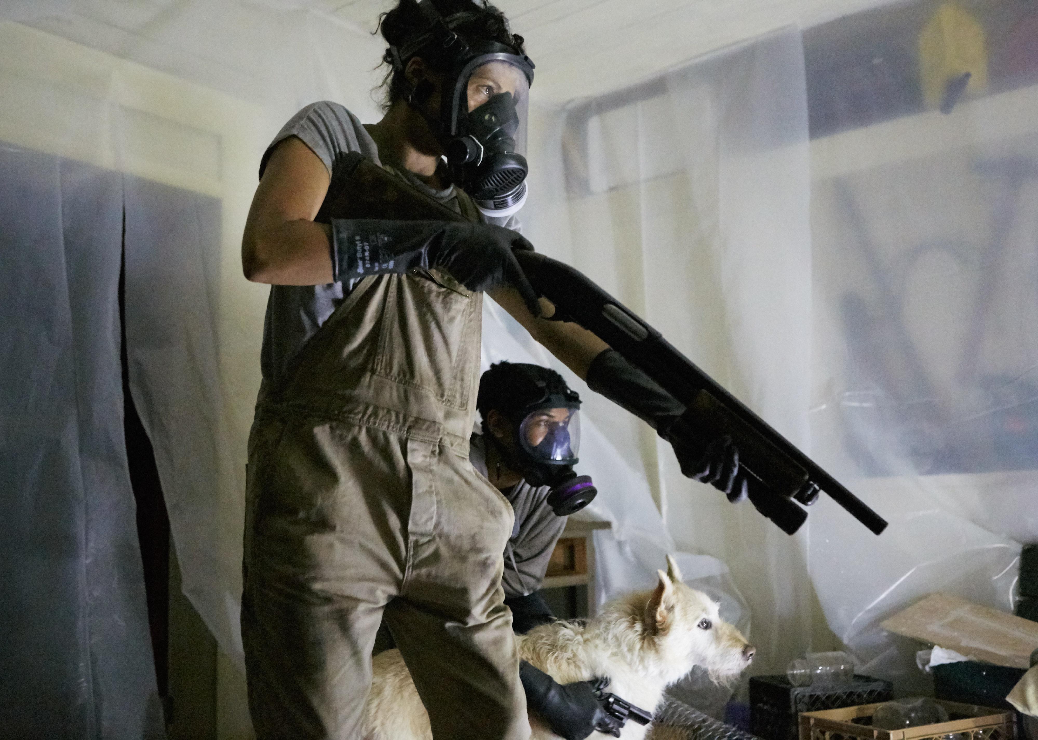 Two women and a dog in a plastic covered room pointing guns while wearing gas masks.